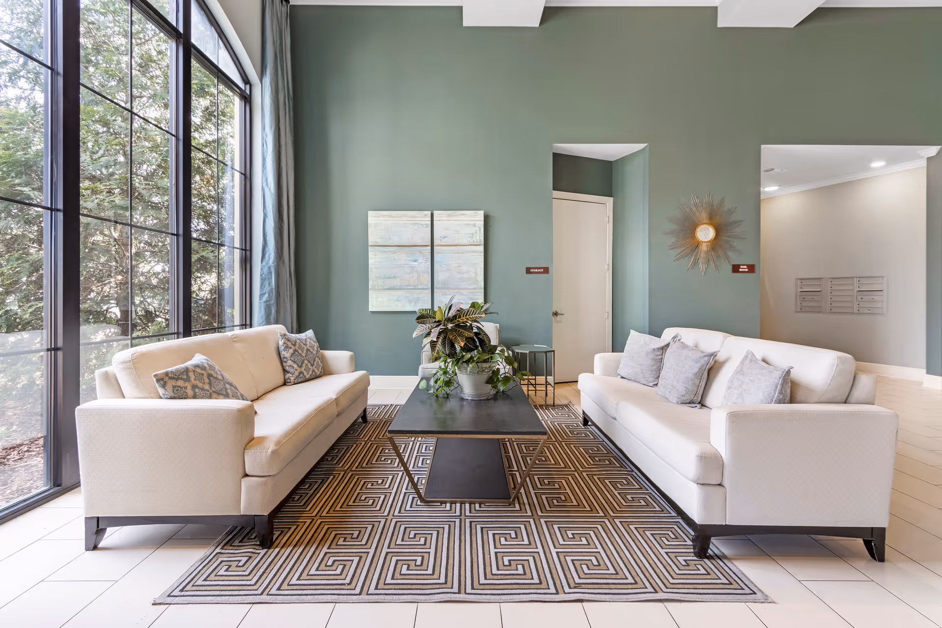 A bright and modern living room with two white sofas facing each other across a black coffee table with a potted plant. The room features a large window with a view of trees outside, green walls, a patterned rug, and decorative wall art including a sunburst mirror.