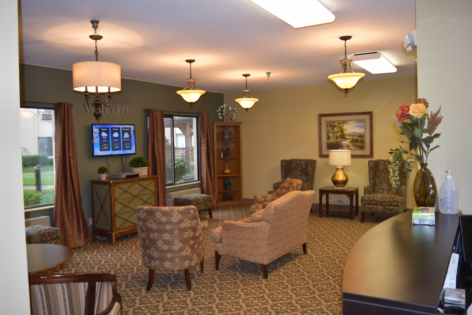 A cozy living room area in Sedgwick Plaza with patterned carpet and multiple upholstered chairs and a loveseat arranged around a small table. The walls are painted in warm tones with framed artwork and a wooden bookshelf. There are three hanging light fixtures on the ceiling, and two windows with brown curtains letting in natural light. A TV mounted on the wall displays a weather forecast. A reception desk with a vase of flowers and hand sanitizer is visible in the foreground.