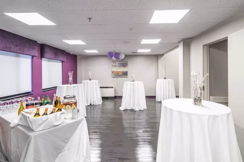 A bright event room with several tall round tables covered in white tablecloths, each decorated with a vase of white branches. One table in the foreground holds a variety of beverages including wine bottles and glasses. The room has purple accent walls and large windows with white blinds.