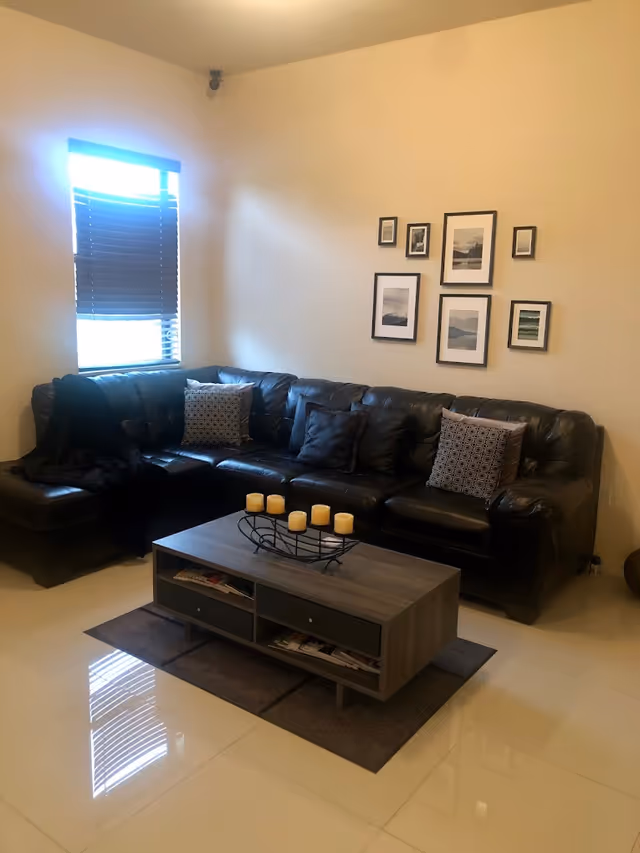 A cozy living room with a black leather sectional sofa adorned with patterned and solid pillows. A wooden coffee table with candles in a decorative holder sits on a rug in front of the sofa. The wall behind the sofa features a collection of framed pictures. A window with closed blinds allows some natural light into the room.
