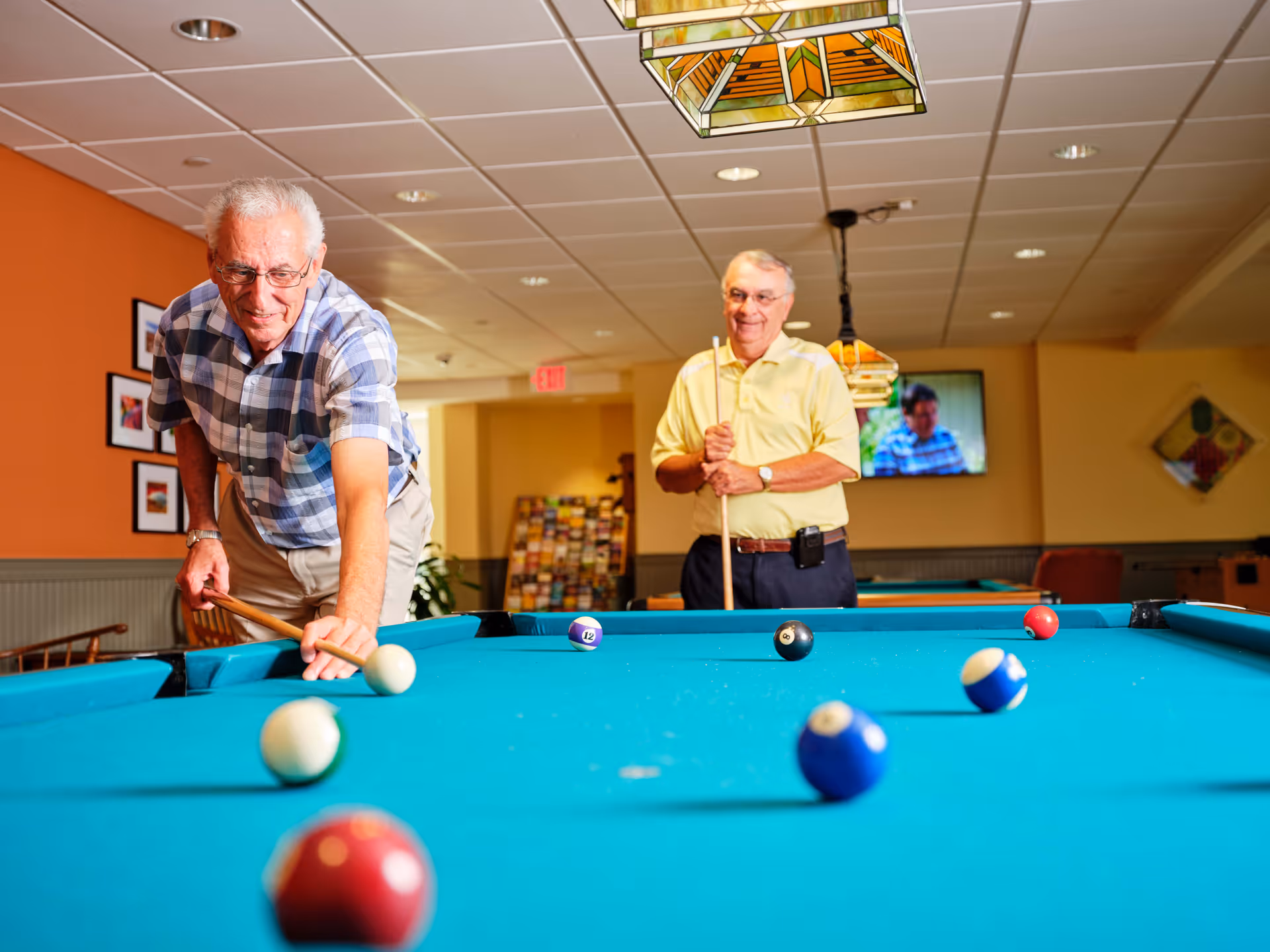 Two elderly men playing pool in a brightly lit recreation room with orange walls and framed pictures. One man is taking a shot while the other watches and smiles, holding a pool cue. There is a television mounted on the wall in the background.