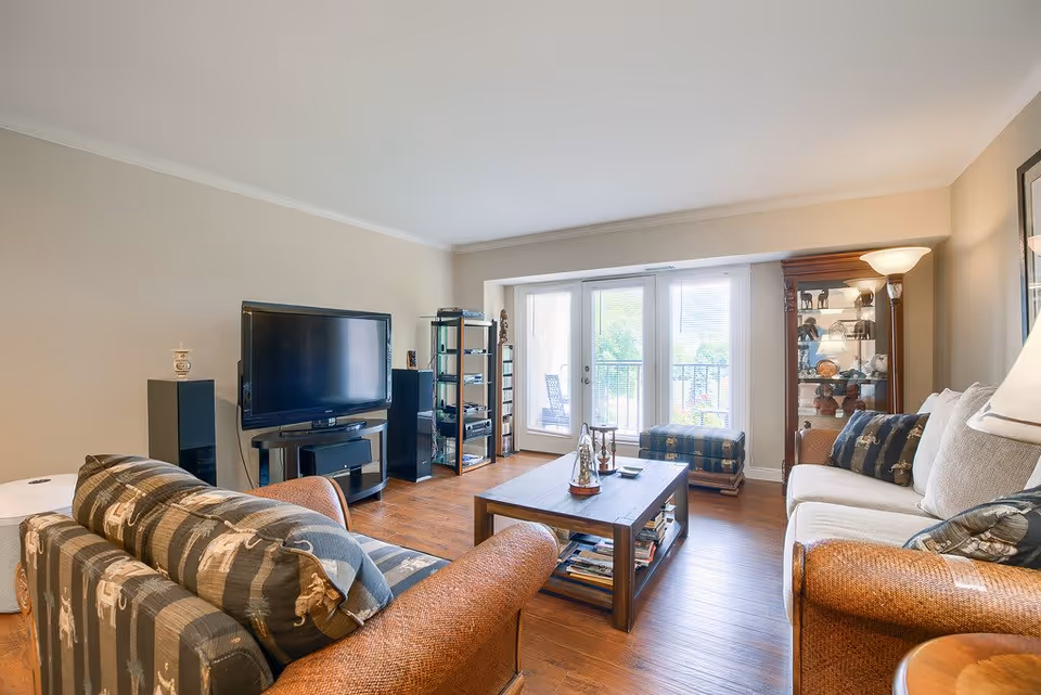 A cozy living room with a large flat-screen TV on a stand, two sofas with patterned cushions, a wooden coffee table with decorative items, a glass display cabinet, and a floor lamp. The room has wooden flooring and large glass doors leading to an outdoor balcony with chairs.