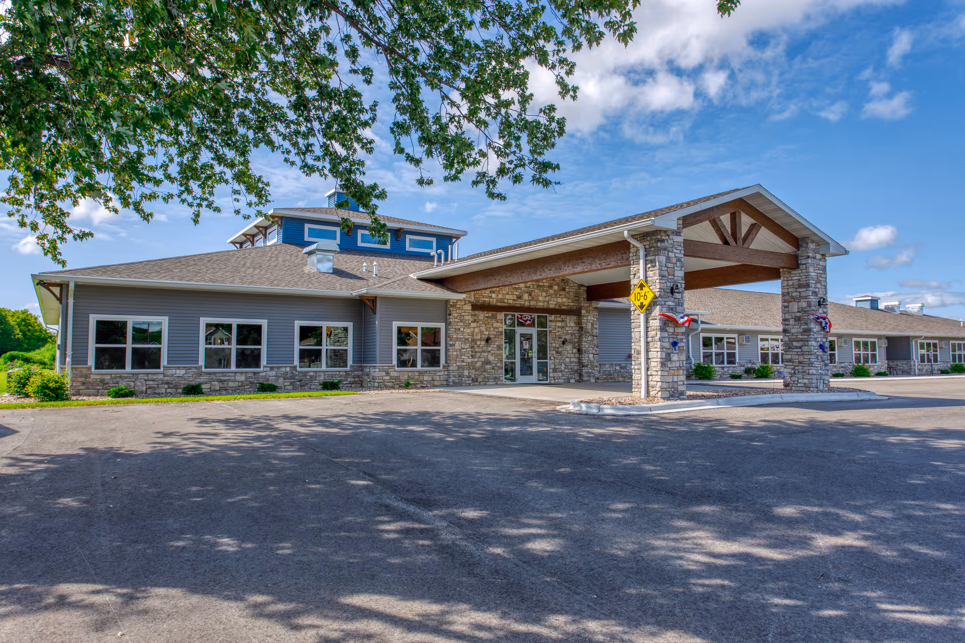 Front exterior of a single-story senior living facility with a stone-clad entrance portico, multiple windows, and a paved driveway under a blue sky.