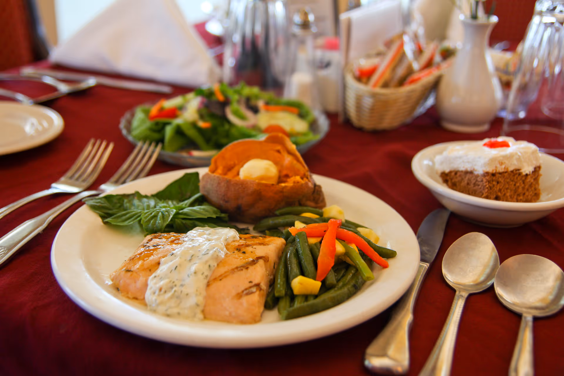 A plated meal on a table with a grilled salmon fillet topped with a creamy dill sauce, steamed mixed vegetables, and a baked sweet potato with butter. In the background, there is a fresh salad and a dessert plate with a slice of cake topped with white frosting and a cherry. The table is set with silverware and a maroon tablecloth.