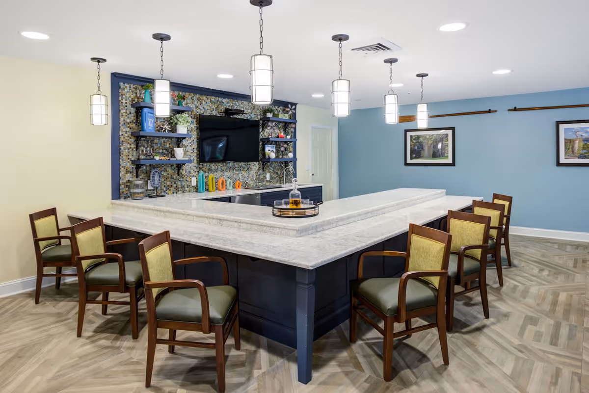 Interior view of a communal dining or gathering area with a large U-shaped marble countertop surrounded by wooden chairs with green cushions. The back wall features a mosaic tile backsplash, open shelves with decorative items, and a mounted flat-screen TV. Pendant lights hang from the ceiling, and framed artwork is displayed on the blue wall to the right.