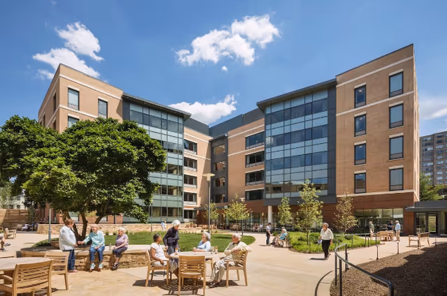 Outdoor courtyard area at Goodwin House Alexandria with several elderly people sitting and standing around wooden tables and benches. The courtyard is surrounded by a modern multi-story brick building with large glass windows. There are trees, small plants, and a clear blue sky with some clouds.