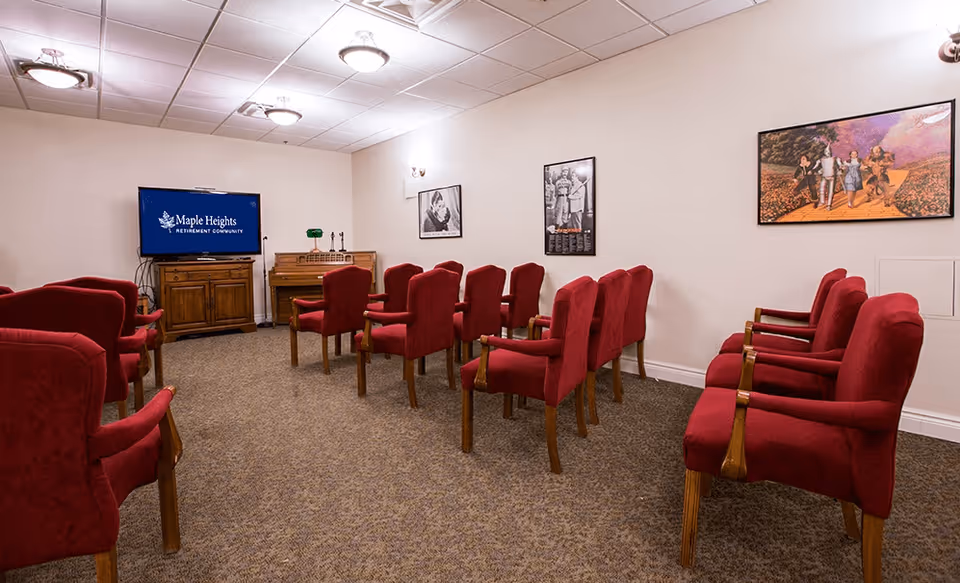 A small theater or presentation room with rows of red upholstered chairs facing a flat-screen TV mounted on a wooden cabinet. The TV screen displays the text 'Maple Heights Retirement Community.' The room has beige walls decorated with framed pictures, a carpeted floor, and ceiling lights.