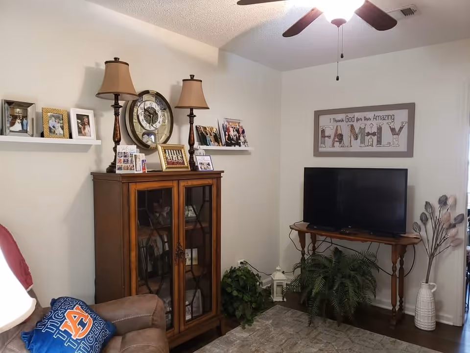A cozy living room corner featuring a wooden cabinet with glass doors, two table lamps, a round wall clock, and several framed family photos on floating shelves. A flat-screen TV sits on a wooden table with plants underneath. A framed sign on the wall reads 'I Thank God for this Amazing FAMILY'. A ceiling fan with lights is visible above, and a brown recliner with a blue Auburn Tigers pillow is partially visible.