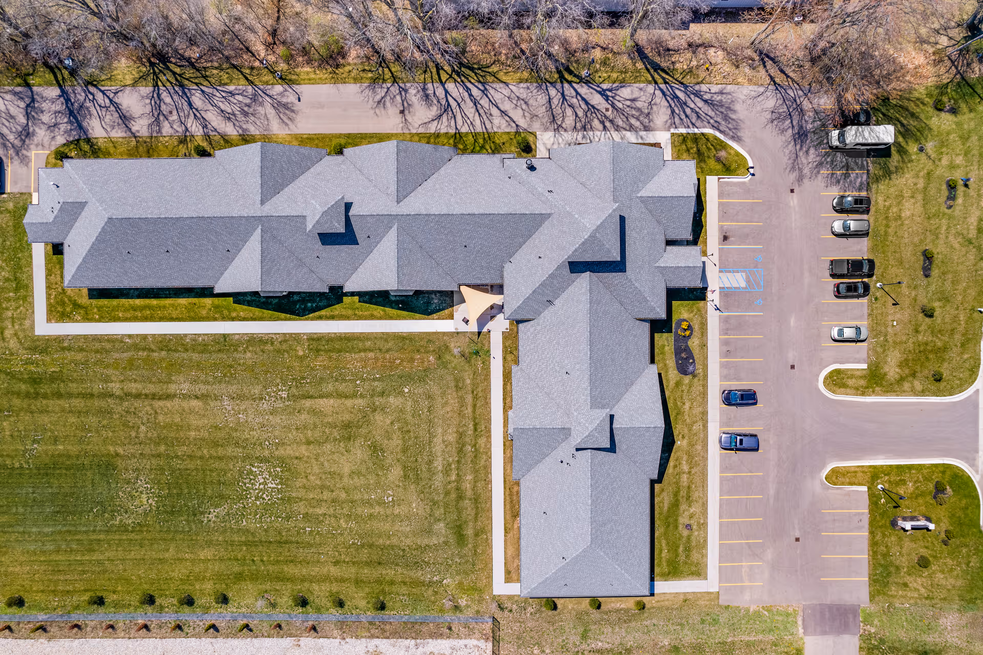 Aerial view of Beacon Pointe Assisted Living facility in Richland showing the building's roof, surrounding green lawn, parking lot with several cars, and a road lined with leafless trees at the top of the image.