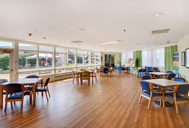 A spacious, well-lit common area with large windows decorated with red, white, and blue star-shaped ornaments. The room has wooden flooring and multiple tables with chairs arranged around them. There are green curtains on one side and a bulletin board and couch in the background.