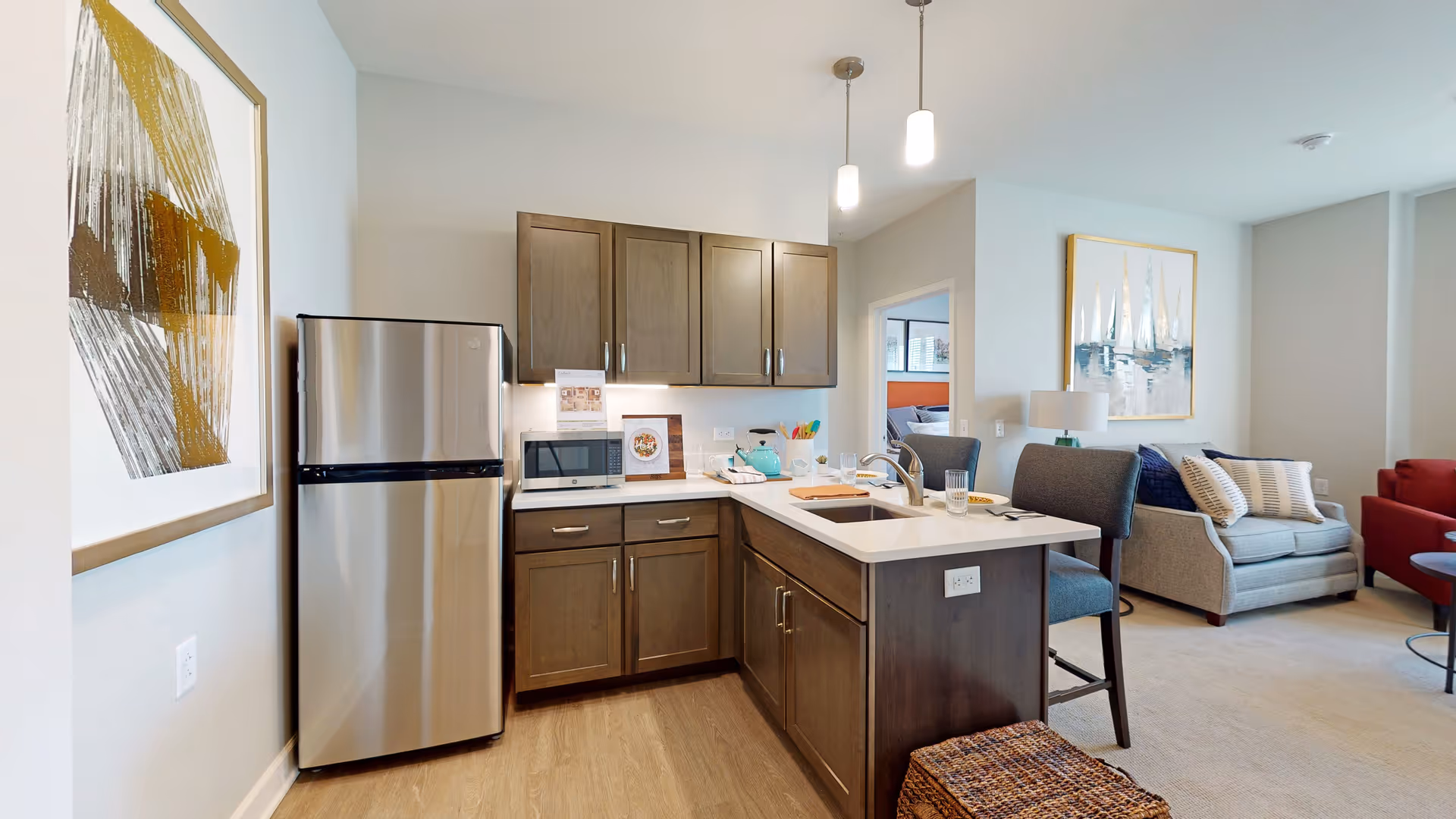 Open-plan kitchen with a stainless refrigerator, wood cabinets and a sink island with barstools opening into a living area with a sofa and artwork.