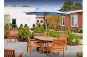 Outdoor patio area with wooden tables and chairs, one table shaded by a large blue umbrella, surrounded by greenery and bushes, with single-story buildings in the background under a clear sky.