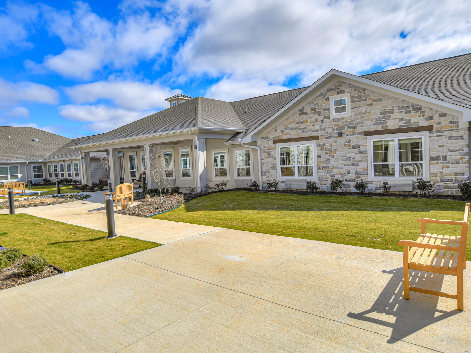Exterior view of a senior living facility building with stone and siding walls, multiple windows, a gray roof, a concrete walkway, green lawn, and wooden benches under a partly cloudy blue sky.