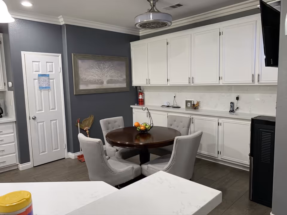 Interior view of a kitchen and dining area with white cabinets, a round wooden table surrounded by four cushioned chairs, a fruit basket on the table, a fire extinguisher on the counter, a broom and dustpan in the corner, and a framed picture of a tree on a dark gray wall.