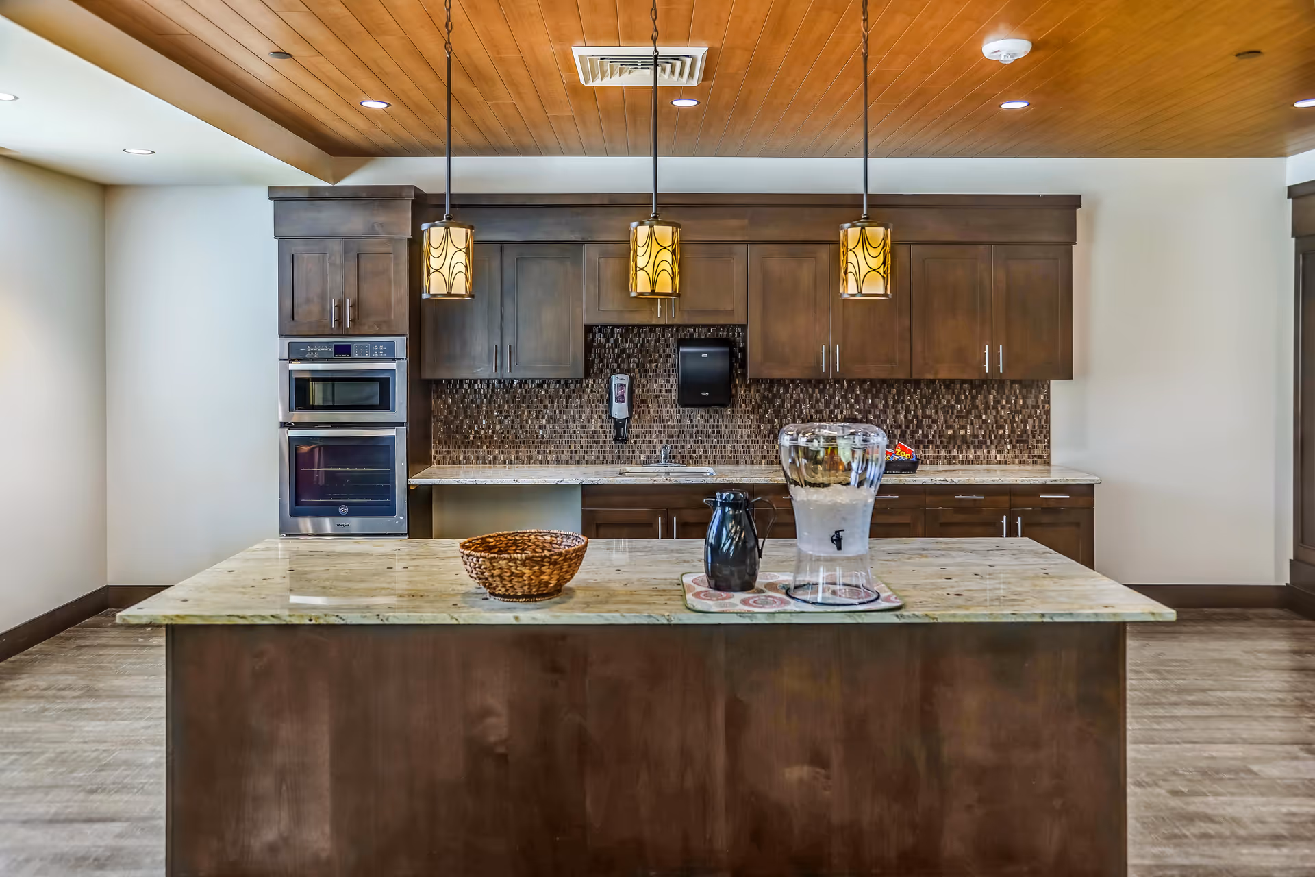 Modern kitchen area with dark wood cabinets, a built-in oven and microwave, a mosaic tile backsplash, and a large island countertop with a basket, a black pitcher, and a glass water dispenser with ice. Three pendant lights hang from the wooden ceiling above the island.