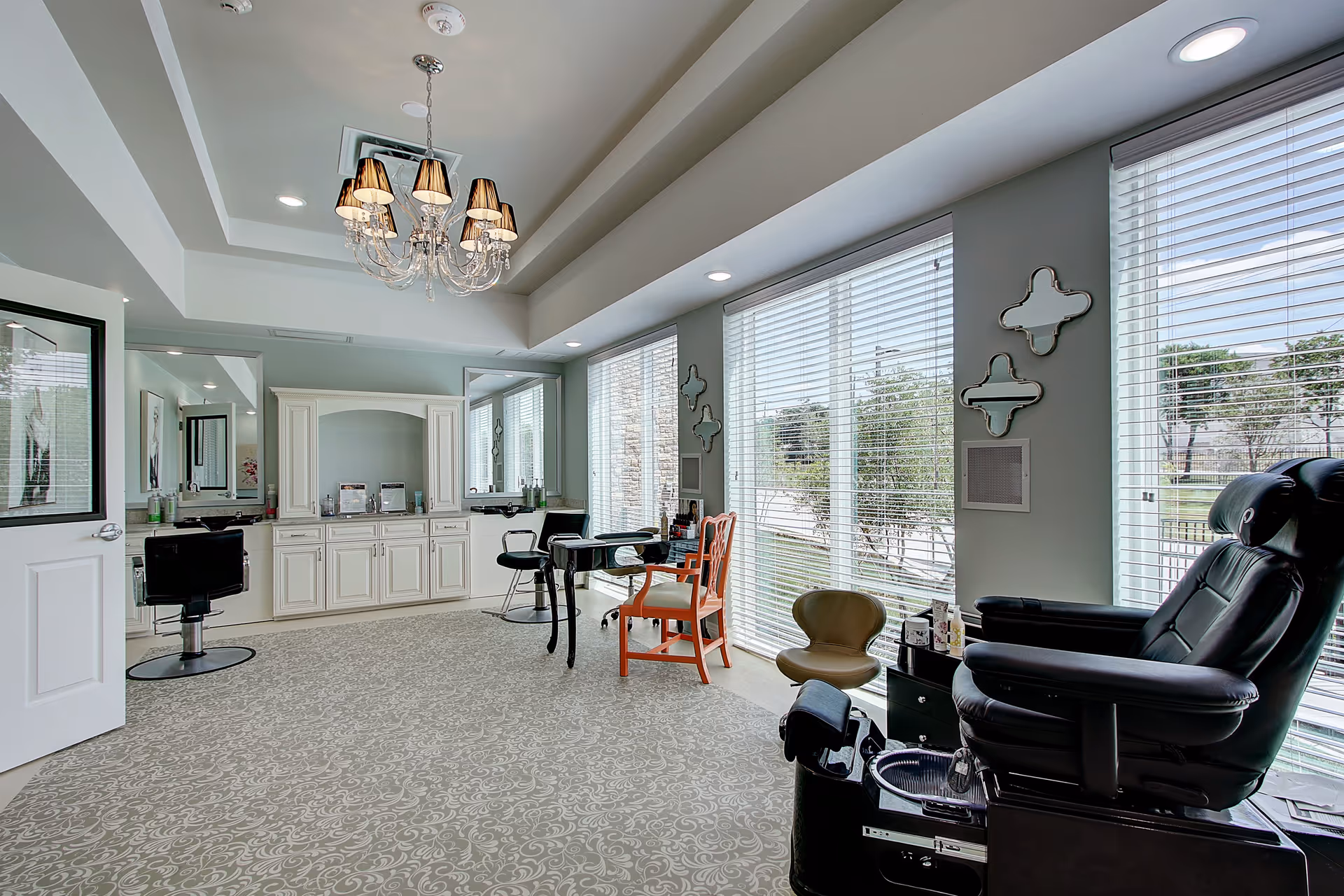 A bright and spacious salon area in The Ridglea Senior Living facility featuring a pedicure chair, styling chairs, a chandelier, large windows with blinds, and decorative wall mirrors.