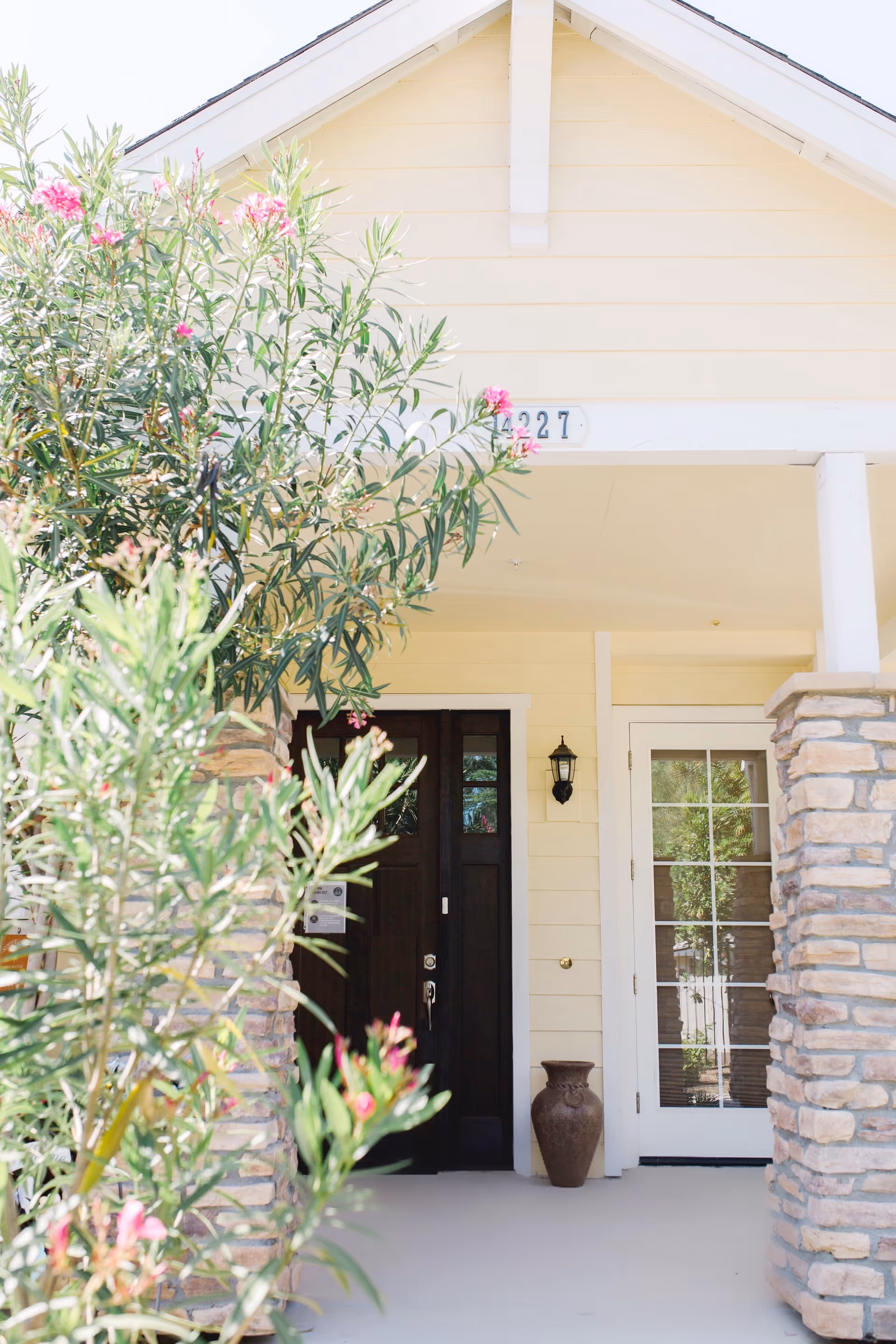 Front entrance of a building with a dark wooden door and a glass-paneled door beside it, flanked by stone pillars and surrounded by green plants with pink flowers.