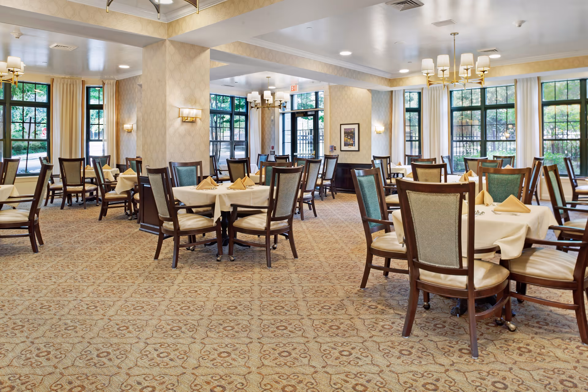 A spacious dining room with multiple tables covered with white tablecloths and beige napkins folded on top. The room features large windows with cream-colored curtains, patterned carpet, and elegant light fixtures including chandeliers and wall sconces. The walls have a subtle patterned wallpaper, and the room is well-lit with natural light coming through the windows.