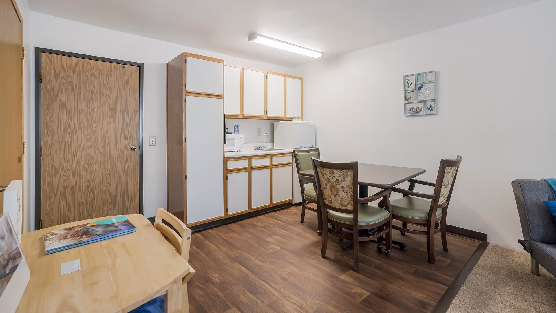 Small dining area with a table and four chairs beside a compact kitchenette with white cabinets and a refrigerator.