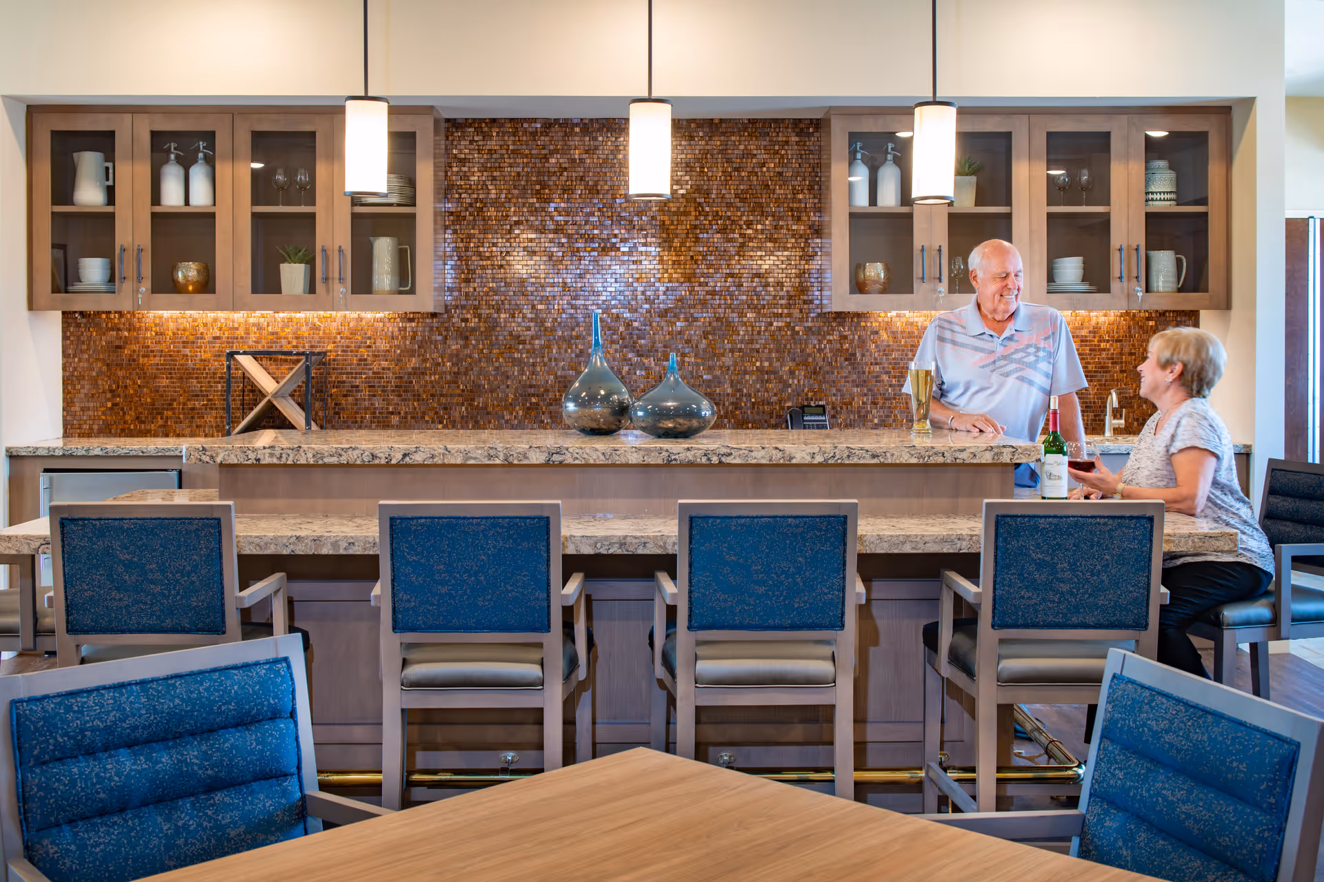 Two people chat at a granite-topped service bar with blue upholstered chairs, pendant lights, and a mosaic tile backsplash in a dining area.