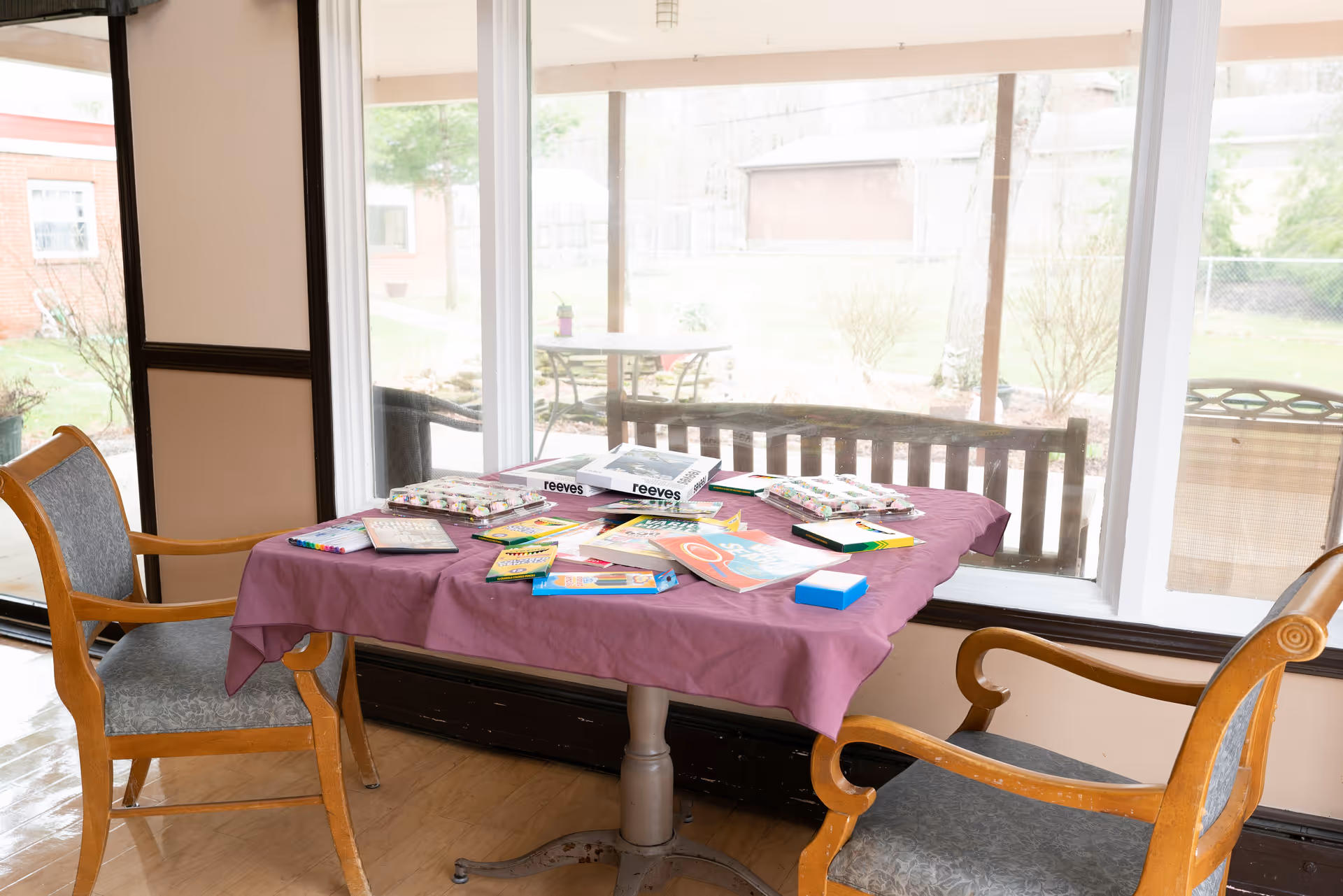 A small table covered with a purple tablecloth is set near large windows overlooking an outdoor area. The table holds various coloring books, crayons, and art supplies. Two wooden chairs with gray cushioned seats are positioned on either side of the table.