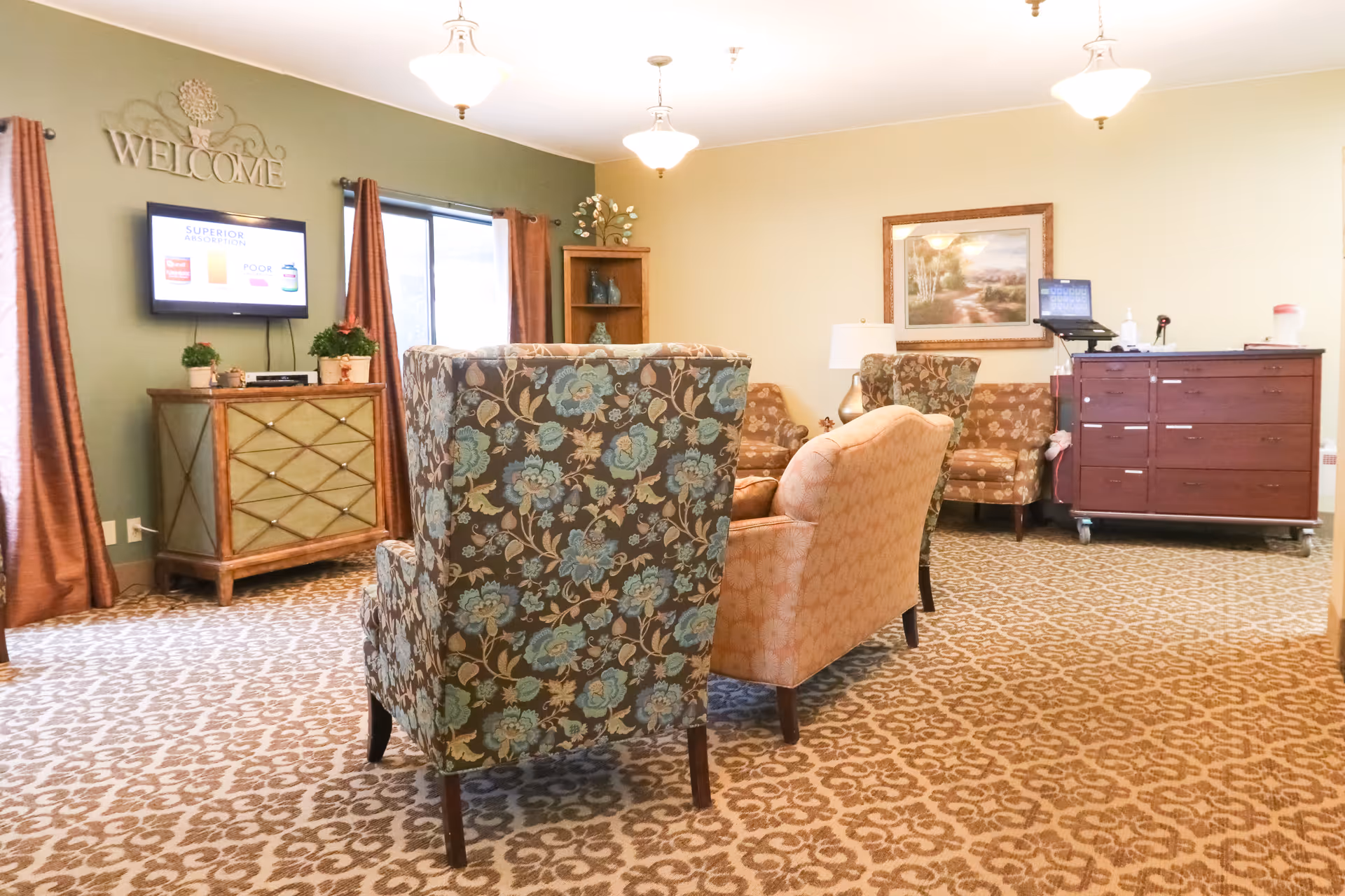 A cozy senior living lounge with patterned armchairs facing a wall-mounted TV, wooden cabinets, and framed artwork under pendant lights.