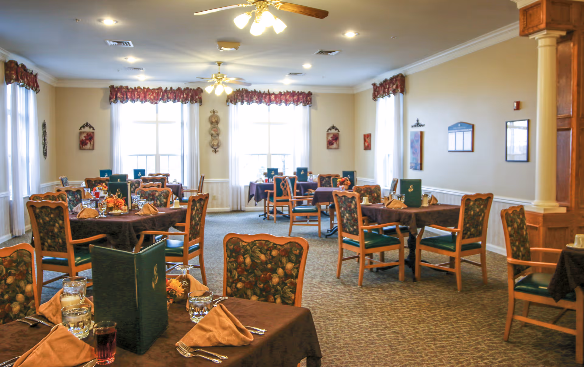 A bright dining room with several tables covered in brown tablecloths, each set with glasses, napkins, and menus. The chairs have wooden frames with floral patterned upholstery. Large windows with white curtains and floral valances let in natural light. Ceiling fans with lights hang from the ceiling, and the walls are decorated with framed artwork and plates.