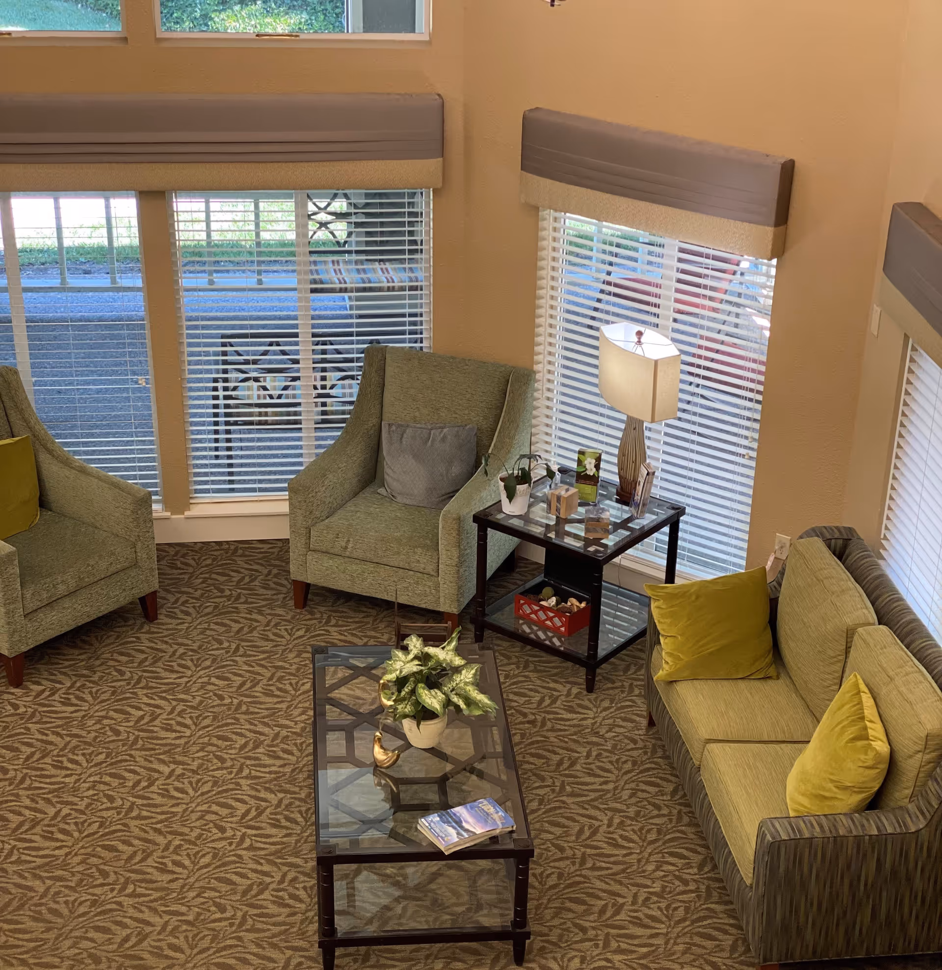 A cozy living room area with two green armchairs and a green sofa with yellow pillows arranged around a glass coffee table with a potted plant and a book on it. There is a side table with a lamp, small decorative items, and brochures next to one of the armchairs. Large windows with blinds allow natural light into the room.