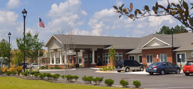 Front exterior of a single-story brick assisted living building with a covered entrance, parked cars, and an American flag.