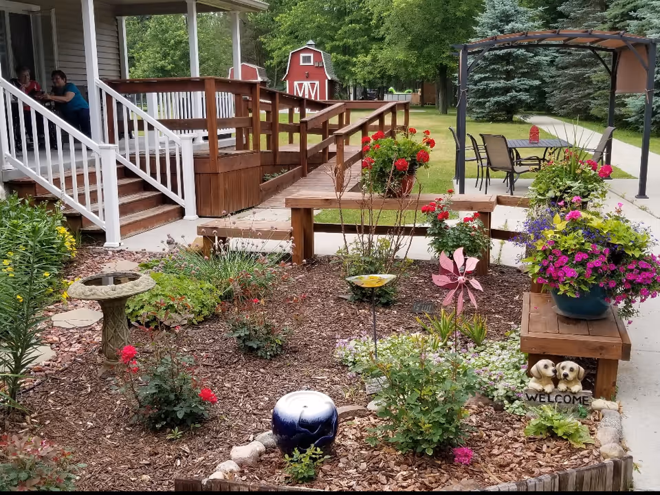 A well-maintained garden area with various colorful flowers and plants, a birdbath, and decorative items including a pink pinwheel and a small statue of two dogs with a welcome sign. There is a wooden ramp and stairs leading to a porch where two people are sitting and talking. In the background, there is a red barn-like structure and a patio area with a table and chairs under a canopy, surrounded by green trees and grass.