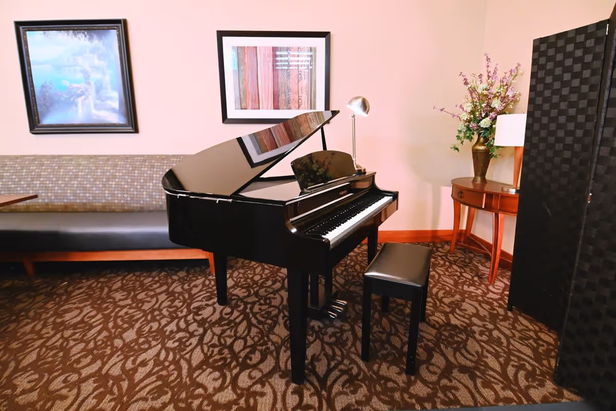 A black grand piano with a matching bench in a room with patterned carpet. Behind the piano is a cushioned bench against the wall with two framed artworks hanging above it. To the right, there is a wooden side table with a lamp and a vase of flowers, and a black folding screen.