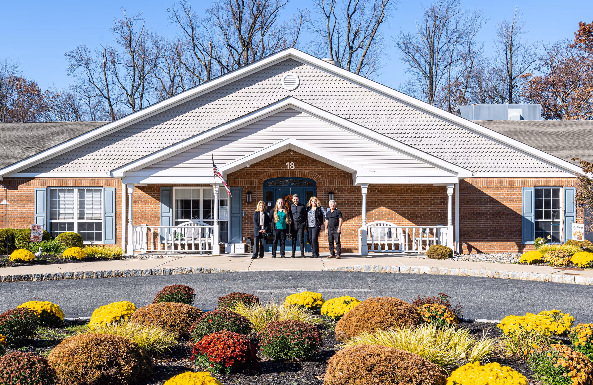 Front exterior view of a single-story brick building with white trim and blue shutters, featuring a covered entrance with white columns and benches. Five people stand in front of the entrance. The foreground has a circular driveway with landscaped bushes and yellow flowers. Trees without leaves are visible in the background under a clear blue sky.