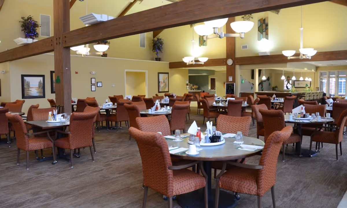 A spacious dining room with multiple round and rectangular tables surrounded by orange upholstered chairs. The tables are set with white napkins, glasses, and condiments. The room features high ceilings with exposed wooden beams, hanging light fixtures, and light yellow walls decorated with framed artwork and potted plants.