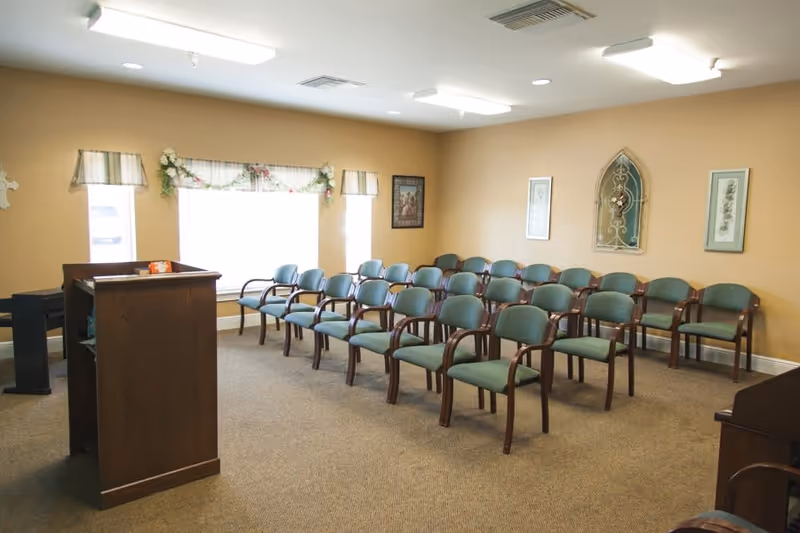 A small meeting or chapel room with rows of green cushioned chairs arranged facing a wooden podium. The room has beige walls with framed artwork and decorative window valances with floral garlands. The floor is carpeted and the room is lit by ceiling lights.