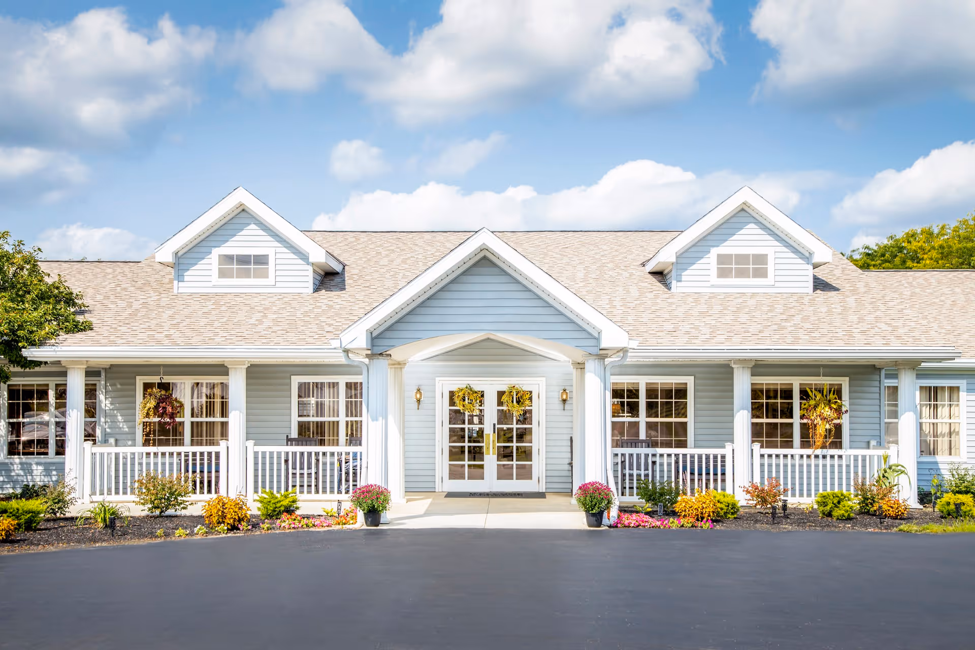 Front exterior view of a light blue senior living facility building with white trim, a covered entrance with double glass doors, white railings on the porch, and colorful flower pots and landscaping in front under a partly cloudy sky.