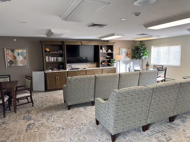 A cozy living room area in a senior living facility featuring two patterned sofas facing a large flat-screen TV mounted on a wooden entertainment center with shelves holding decorative items and books. There is a dining table with chairs to the left, a large window with blinds on the right, and a tall potted plant near the window. The room has neutral-colored walls and carpeted flooring with a subtle pattern.