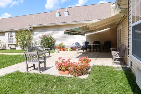 Outdoor patio area at Vintage Park at Ottawa with wooden benches, a table with chairs under a retractable awning, potted plants, and green grass surrounding the paved patio.