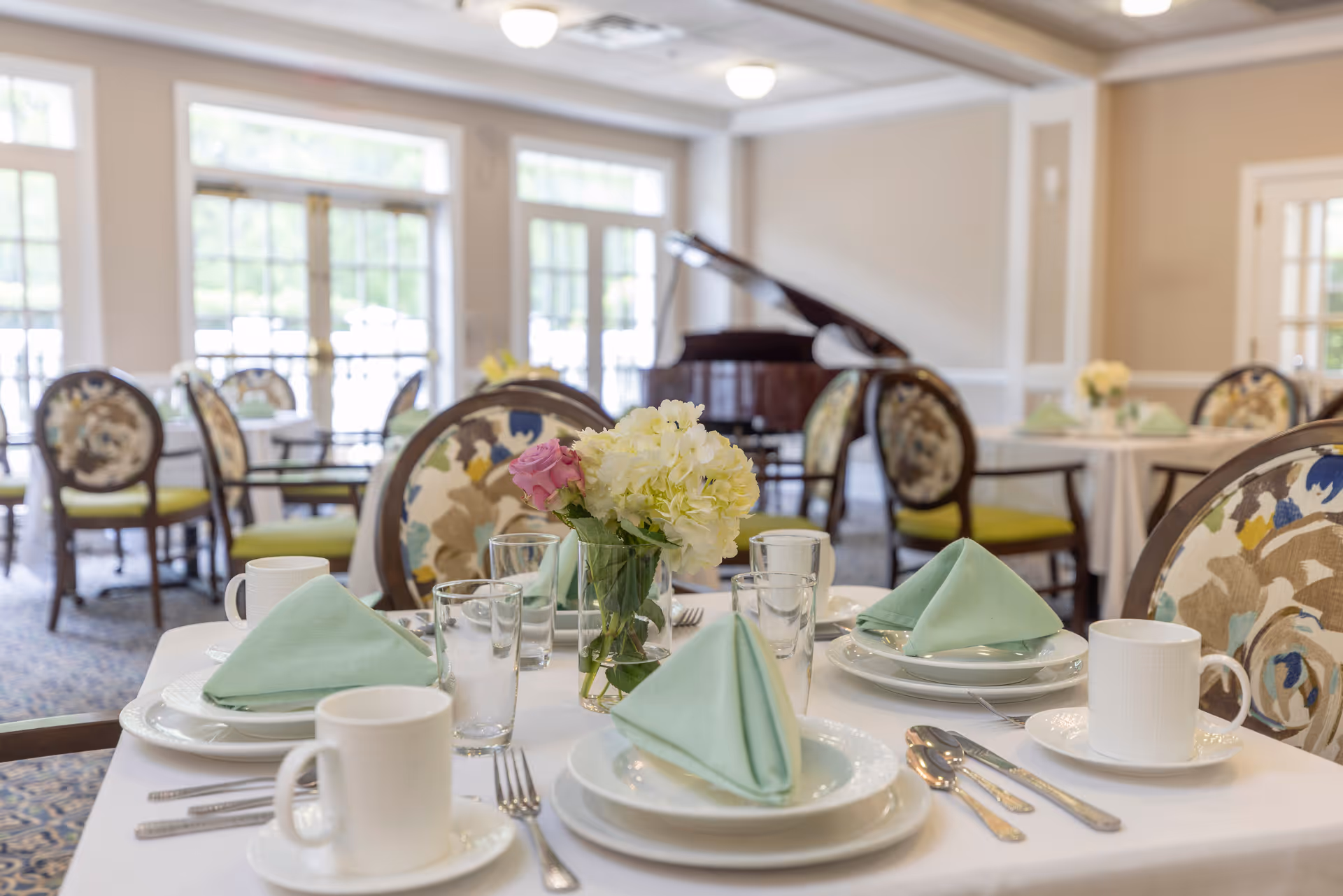 A dining room set up with tables covered in white tablecloths, each table featuring neatly folded green napkins, white plates, cups, glasses, and silverware. A small vase with white and pink flowers is placed in the center of the table. The room has large windows allowing natural light to fill the space, and a grand piano is visible in the background.