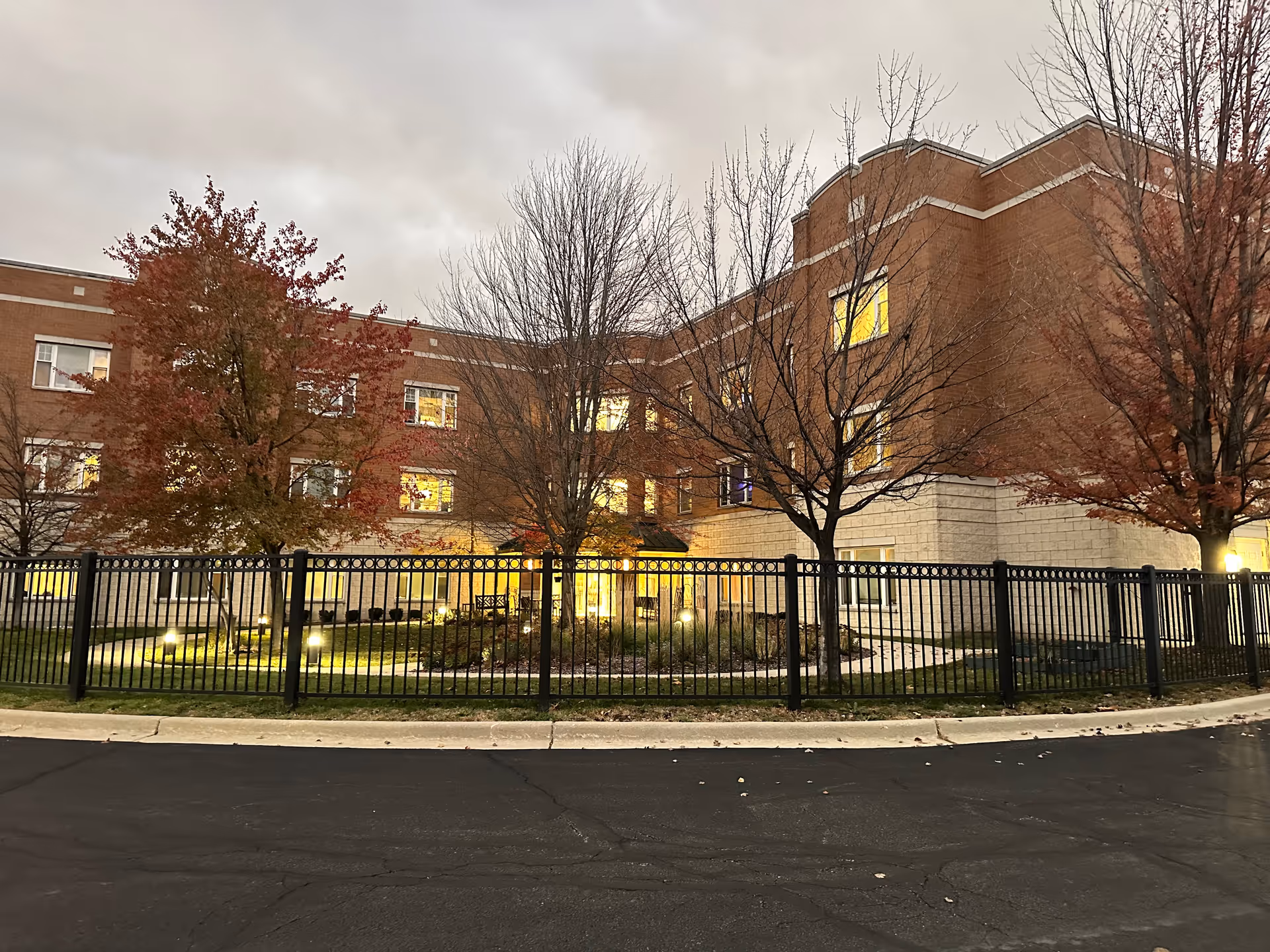 Exterior view of a multi-story brick building with lit windows during dusk. The building is surrounded by a black metal fence and several leafless and autumn-colored trees. There is a small landscaped garden area with lights inside the fenced area.