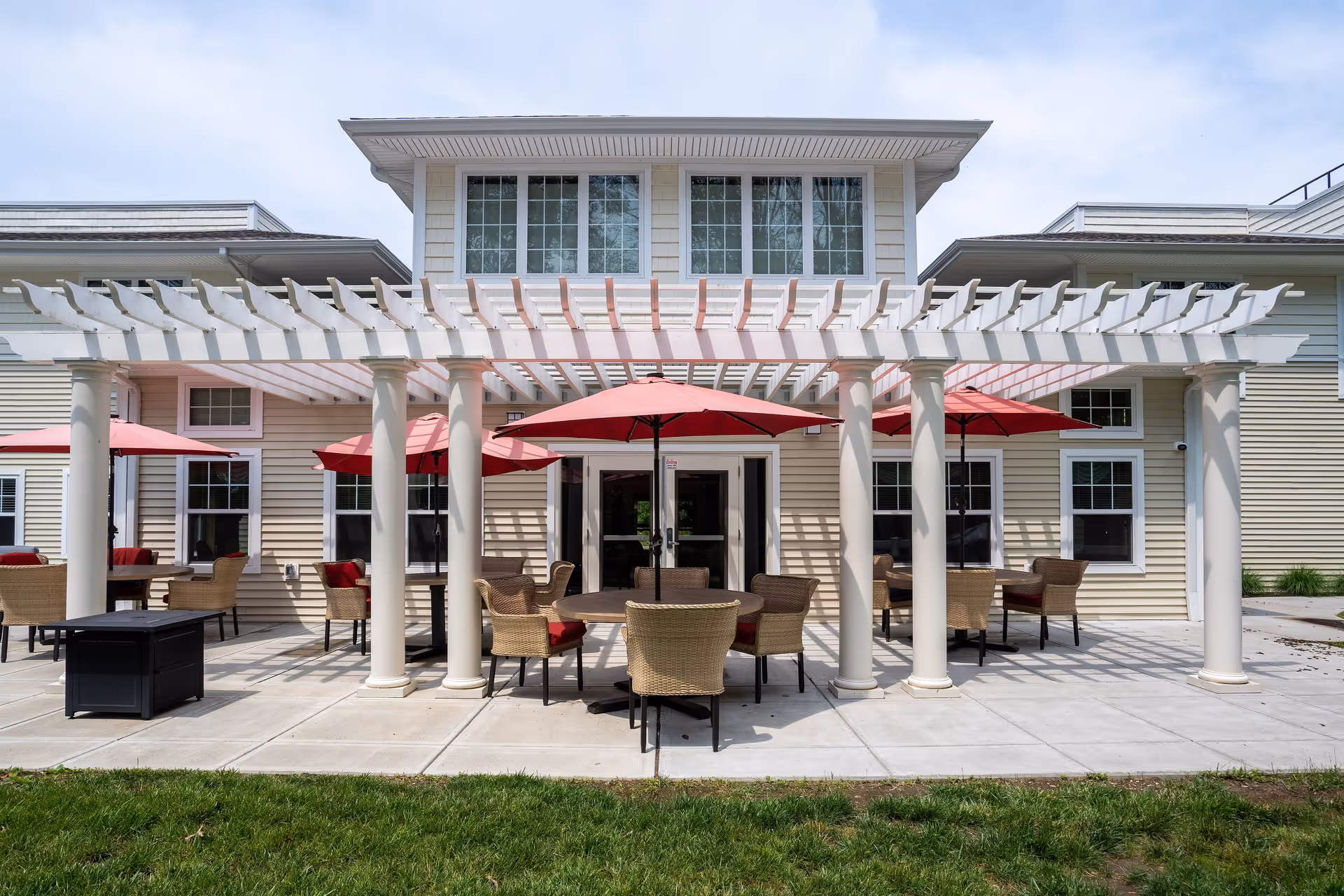 Outdoor patio with round tables, wicker chairs and red umbrellas under a white pergola in front of a two-story building.