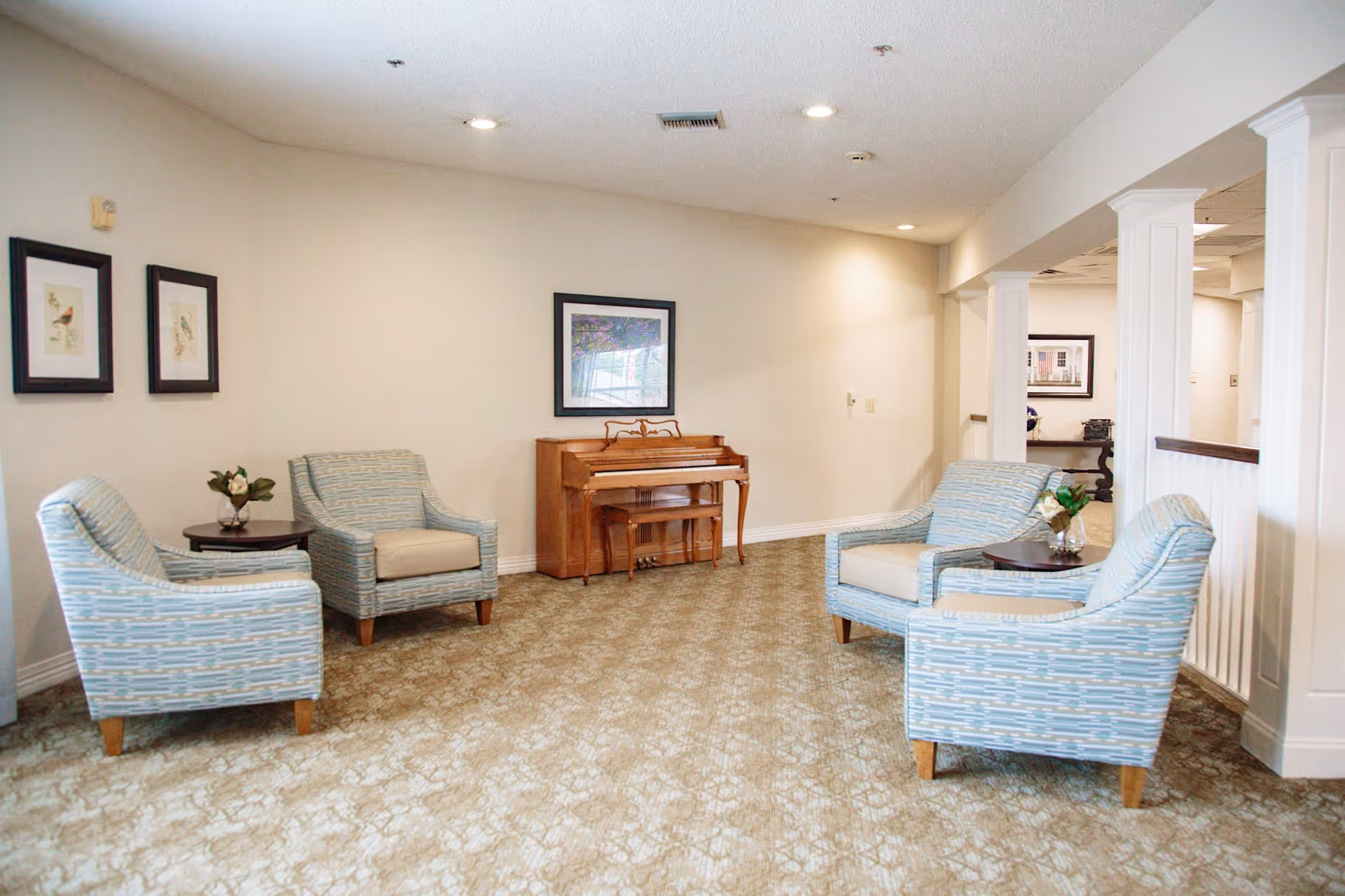 A cozy sitting area with four light blue patterned armchairs arranged around two small round tables with flower vases. A wooden piano with a matching bench is against the wall, which is decorated with framed artwork. The room has beige walls, a patterned carpet, and recessed ceiling lights.