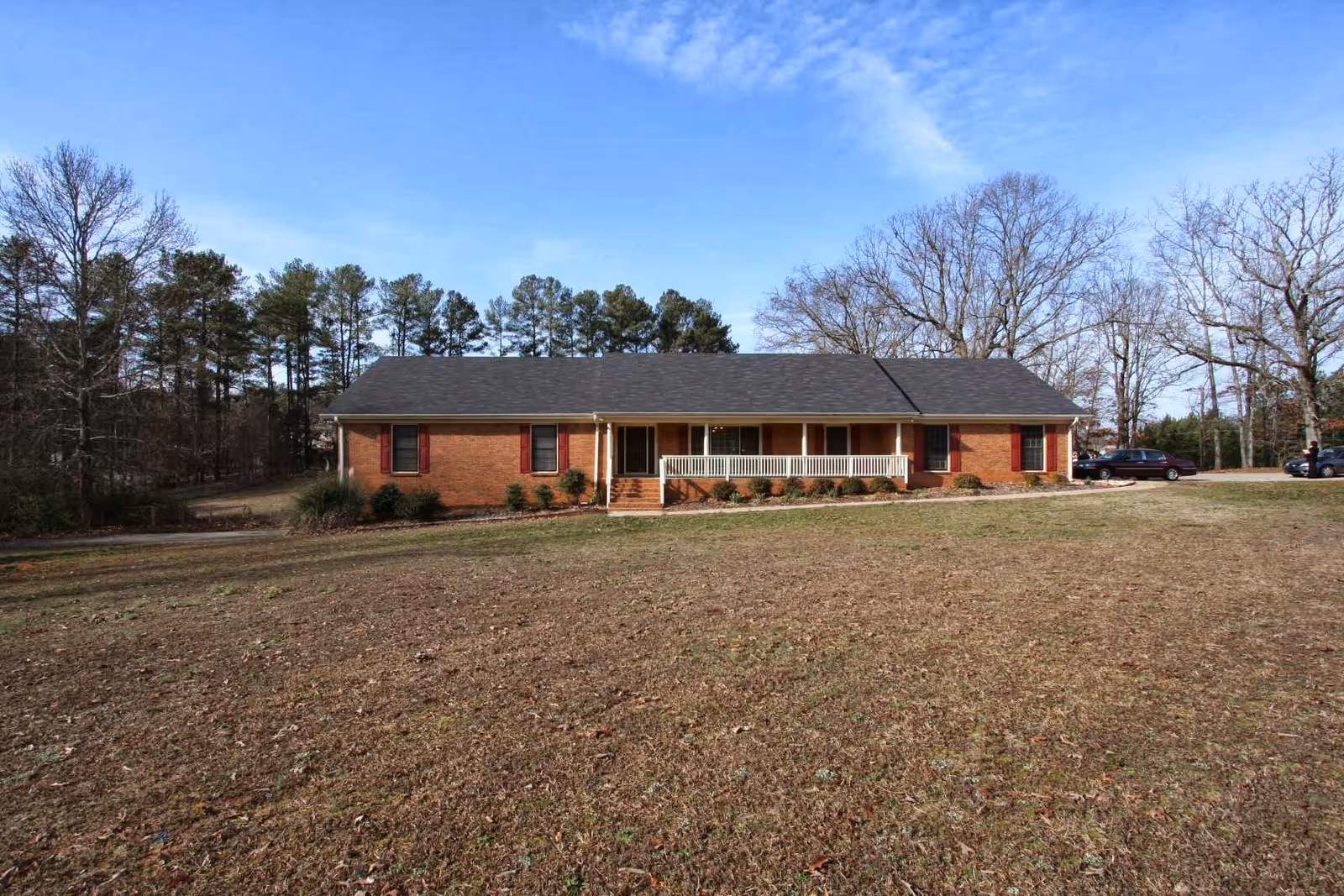 Single-story brick building with a dark shingled roof, a front porch with white railing, and several windows with red shutters. The building is surrounded by a large grassy area with some trees in the background under a blue sky with light clouds.