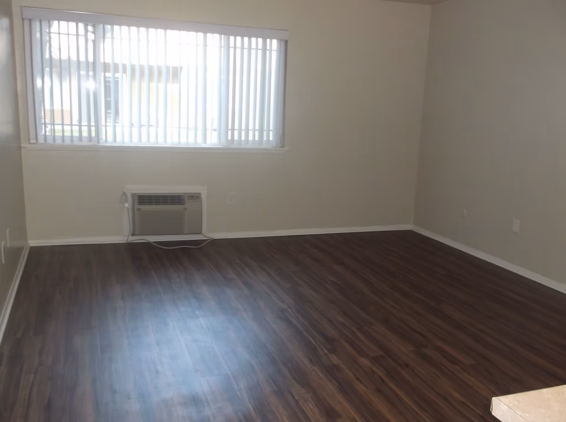 Empty room with dark wood flooring, beige walls, a large window with vertical blinds, and a wall-mounted air conditioning unit below the window.