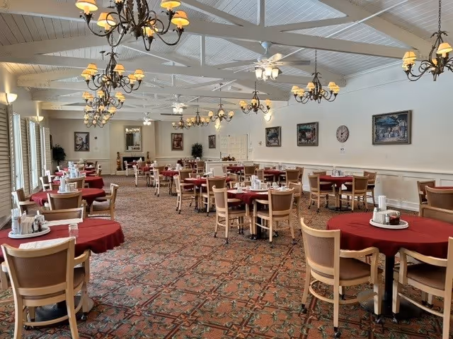 Spacious dining room with round tables covered in red tablecloths, wooden chairs, chandeliers, and patterned carpet.
