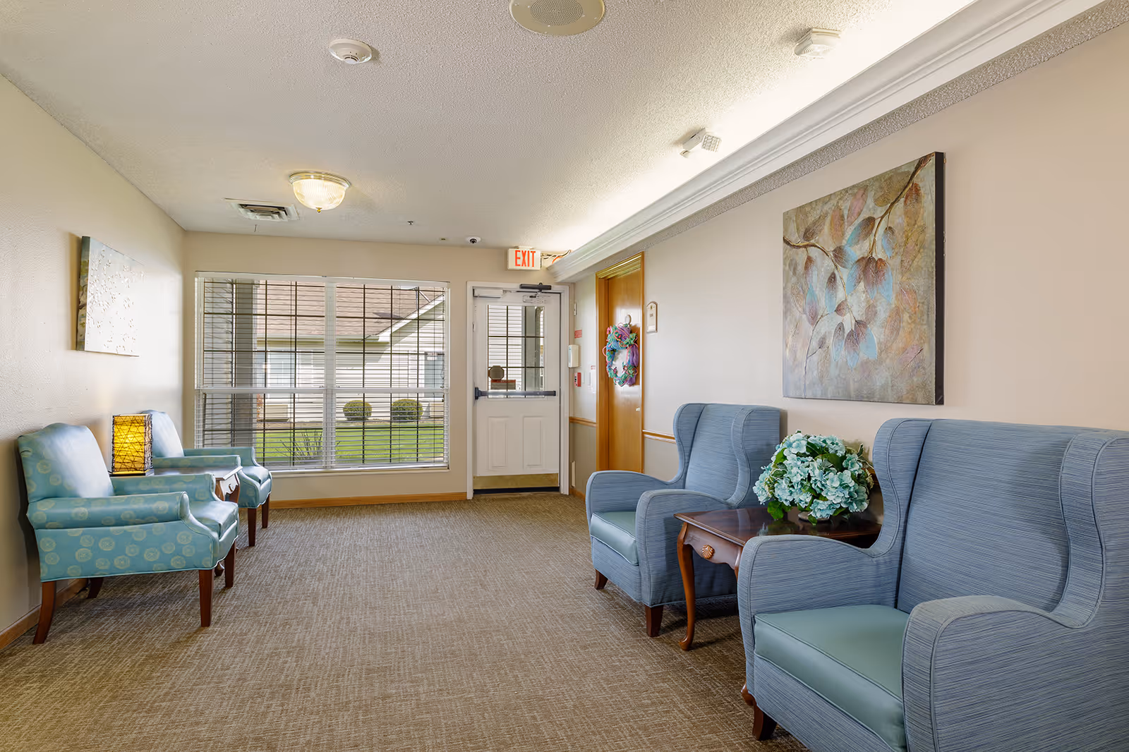 A bright and clean hallway seating area in an assisted living facility featuring two pairs of upholstered armchairs in blue tones, a small wooden side table with a floral arrangement, a large window with blinds, a door with an exit sign above it, and a decorative painting on the wall.