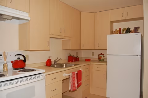 A compact kitchen with light wood cabinets, a white refrigerator, a white stove with a red kettle on it, a stainless steel sink, and various red kitchen accessories including towels, containers, and a decorative jar. The word 'HOME' is displayed on top of the refrigerator.