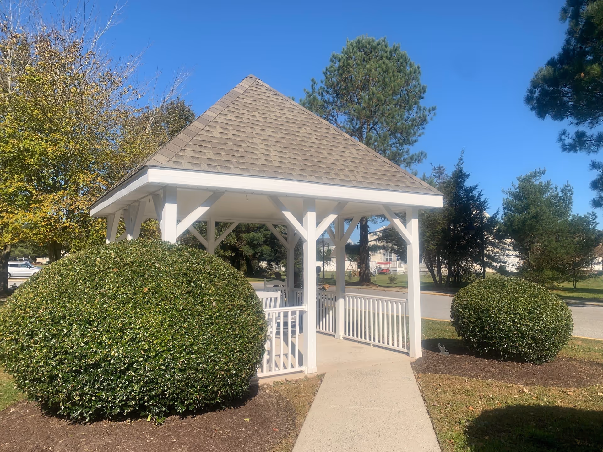 A small white gazebo with a shingled roof situated on a paved walkway, surrounded by neatly trimmed bushes and trees under a clear blue sky.