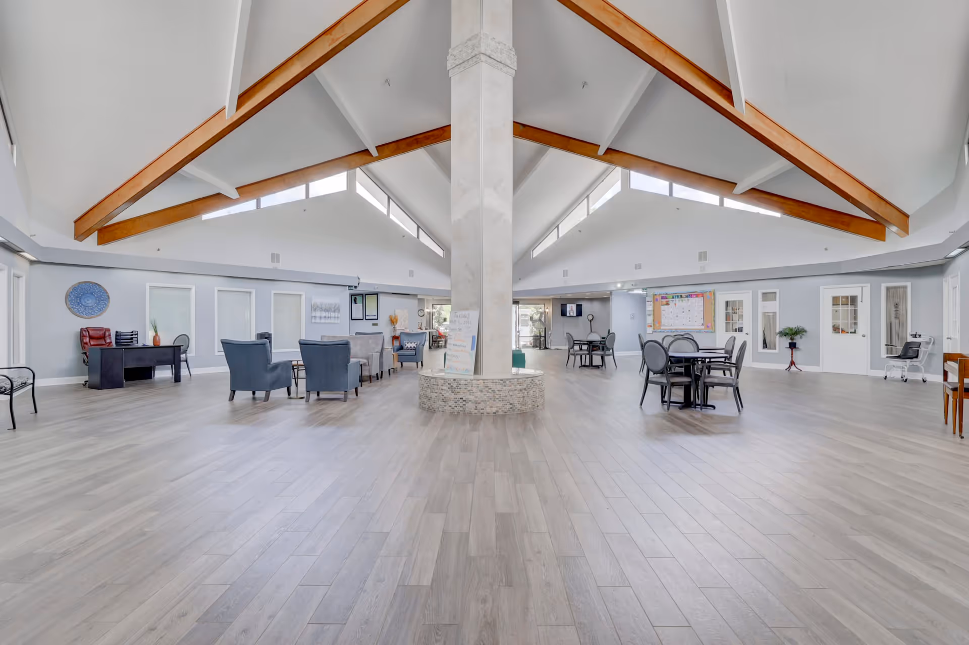Spacious senior living facility common area with high vaulted ceilings featuring wooden beams, light gray walls, and wood-look flooring. The room is furnished with several seating areas including armchairs, sofas, and round tables with chairs. A central column is decorated with mosaic tiles at the base and has a whiteboard attached. Large windows near the ceiling allow natural light to fill the space.