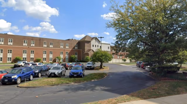 Front exterior of a multi-story brick senior living building with a parking lot, driveway, trees, and a partly cloudy sky.