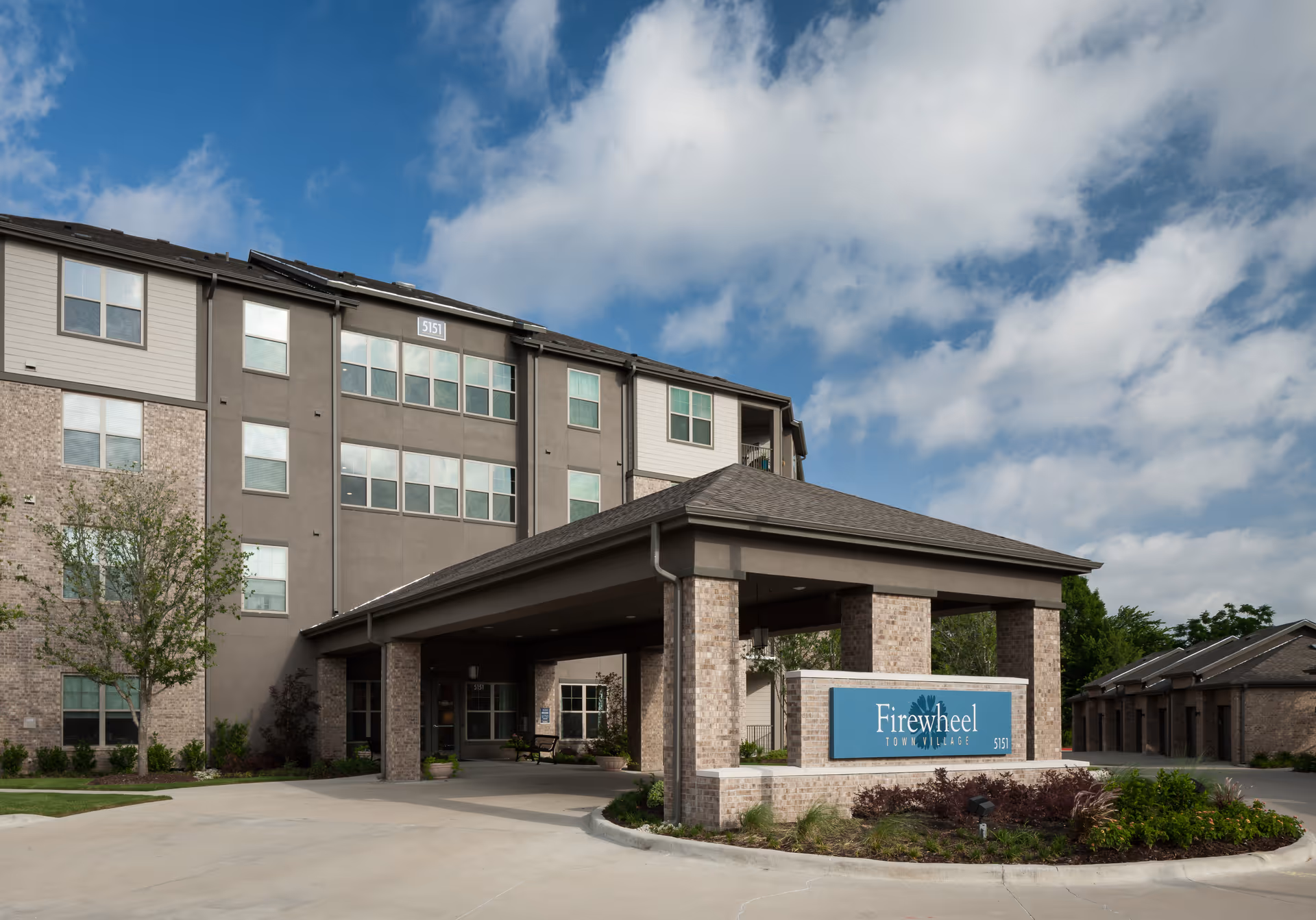Exterior view of Firewheel Town Village building with a covered entrance supported by brick columns, a sign displaying the facility name, and a partly cloudy blue sky above.
